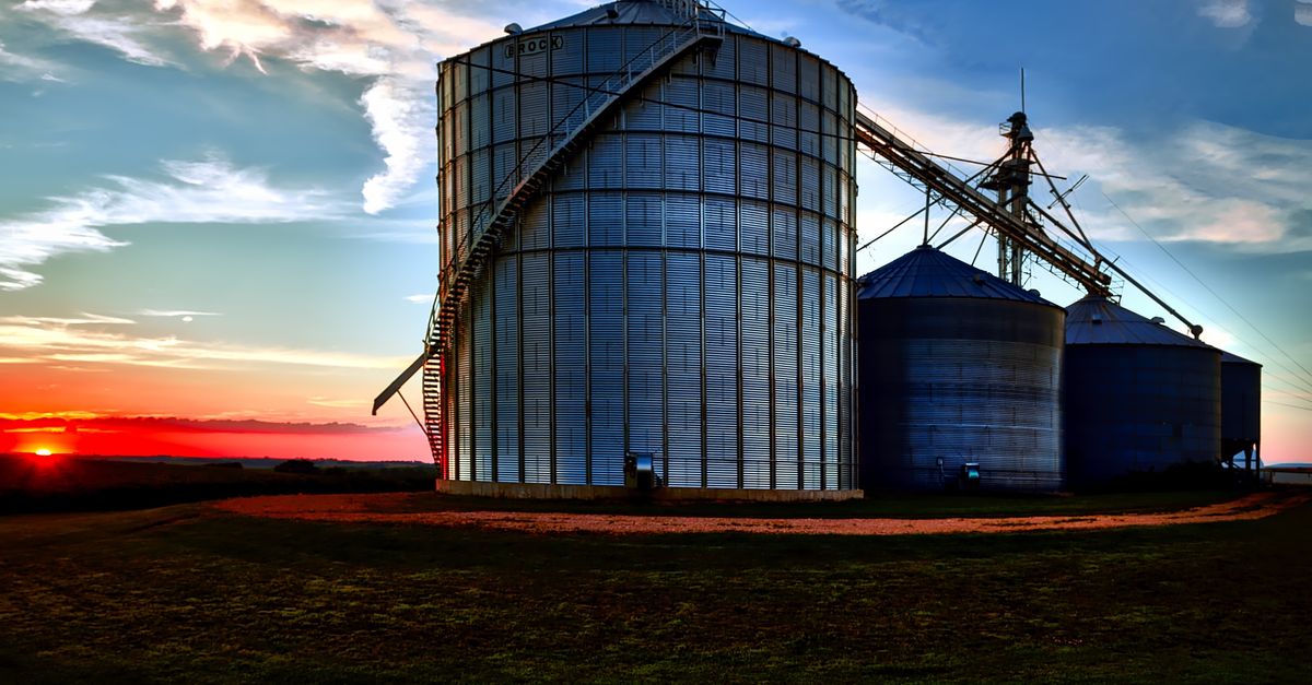Large grain silos under a dramatic sunset sky, representing agriculture and industry in a rural setting.