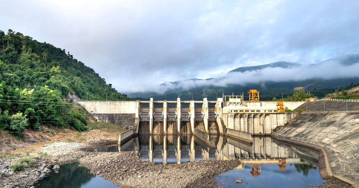 Industrial hydroelectric power plant located near river and stony coast against green mountain range covered with mist o